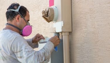 Professional house painter wearing protective respirator while masking an electrical meter.